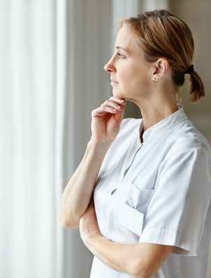 Nurse in a white uniform standing by a window, looking thoughtful