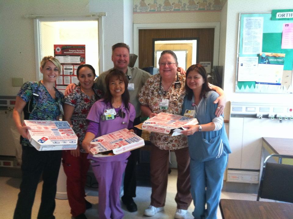 Six nurses in scrubs holding open pizza boxes in a break room