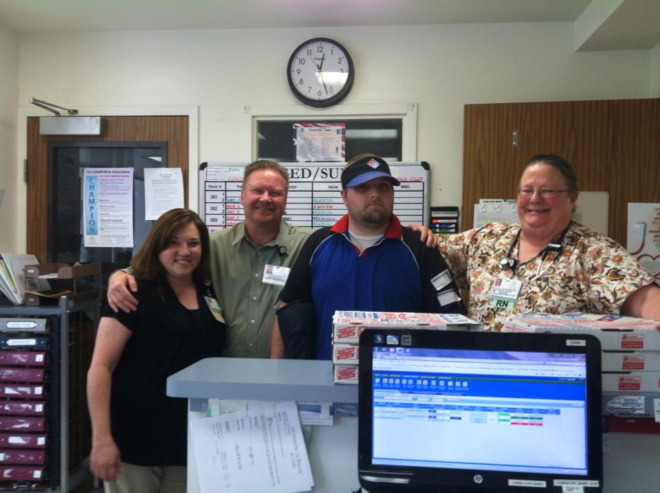 Four hospital staff smiling behind a nurses station counter stacked with pizza boxes
