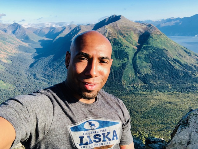 Selfie in an Alaska T-shirt with green mountains and water behind