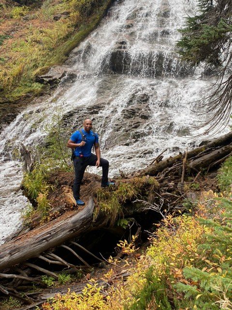 Hiker in a blue jacket crossing a log at the foot of a waterfall