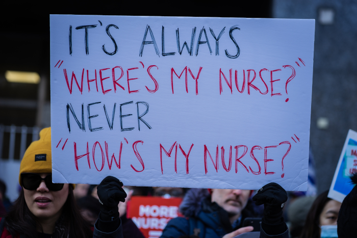 Protesters hold a hand-lettered sign: it’s always where’s my nurse, never how’s my nurse