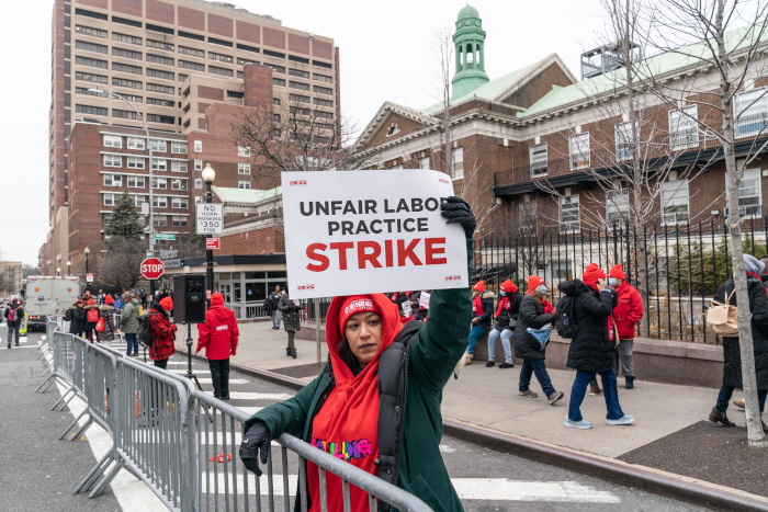 Nurse in a red hat on a city picket line holding an Unfair Labor Practice Strike sign