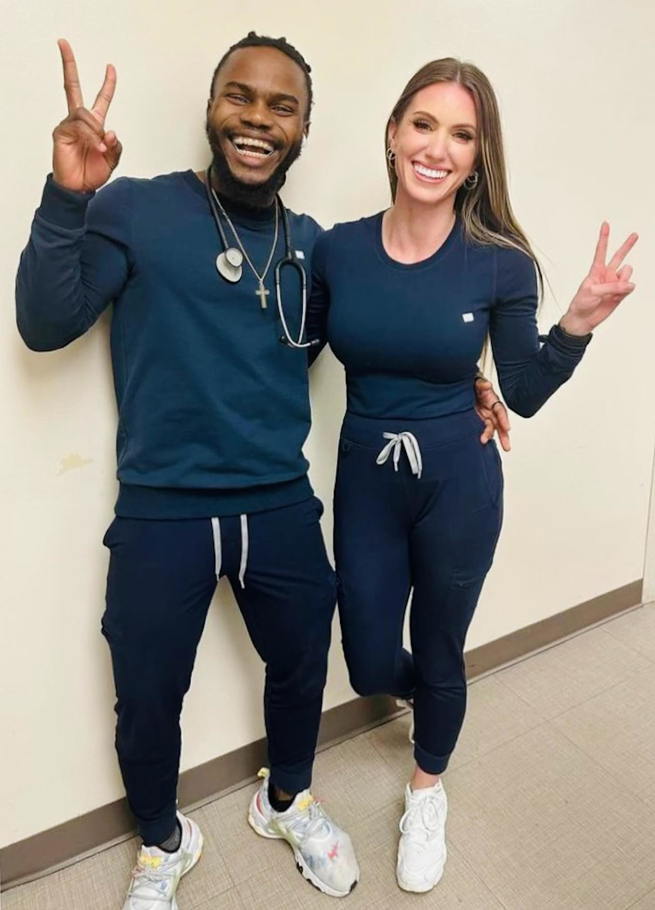 Two nurses in navy scrubs grinning and flashing peace signs against a hallway wall