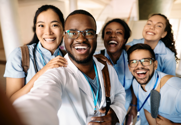 Five smiling healthcare workers in scrubs crowding into a group selfie